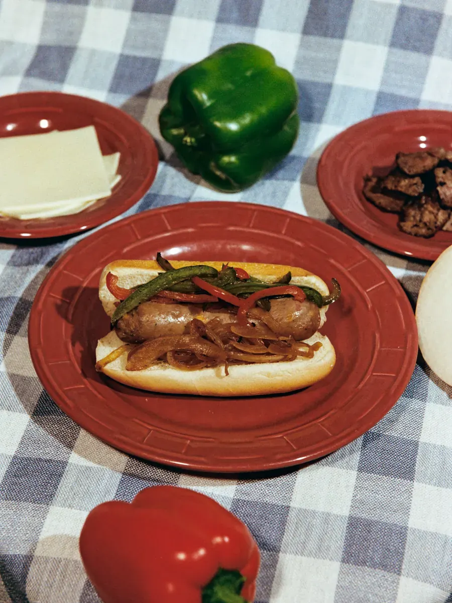 PhillyWürst on a bun with peppers and onions, beside bell peppers, provolone and steak
