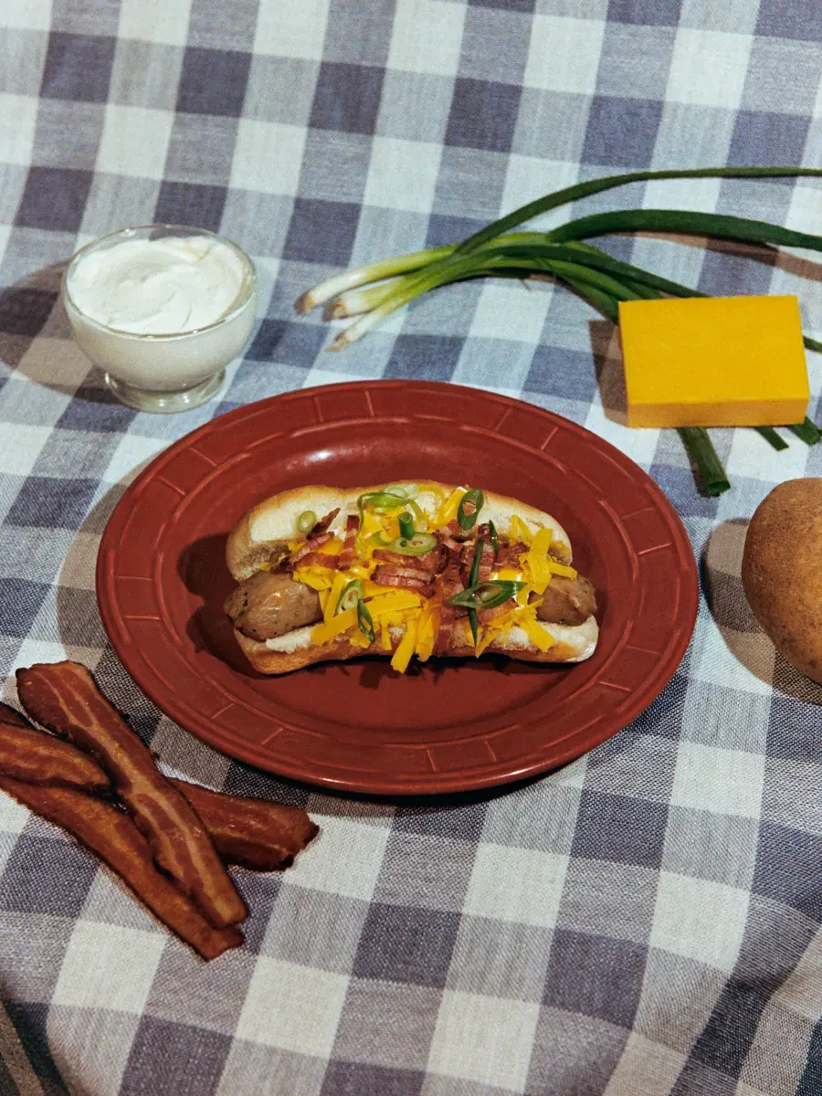 TaterWürst on a bun with cheddar, bacon and scallion, beside sour cream and a potato